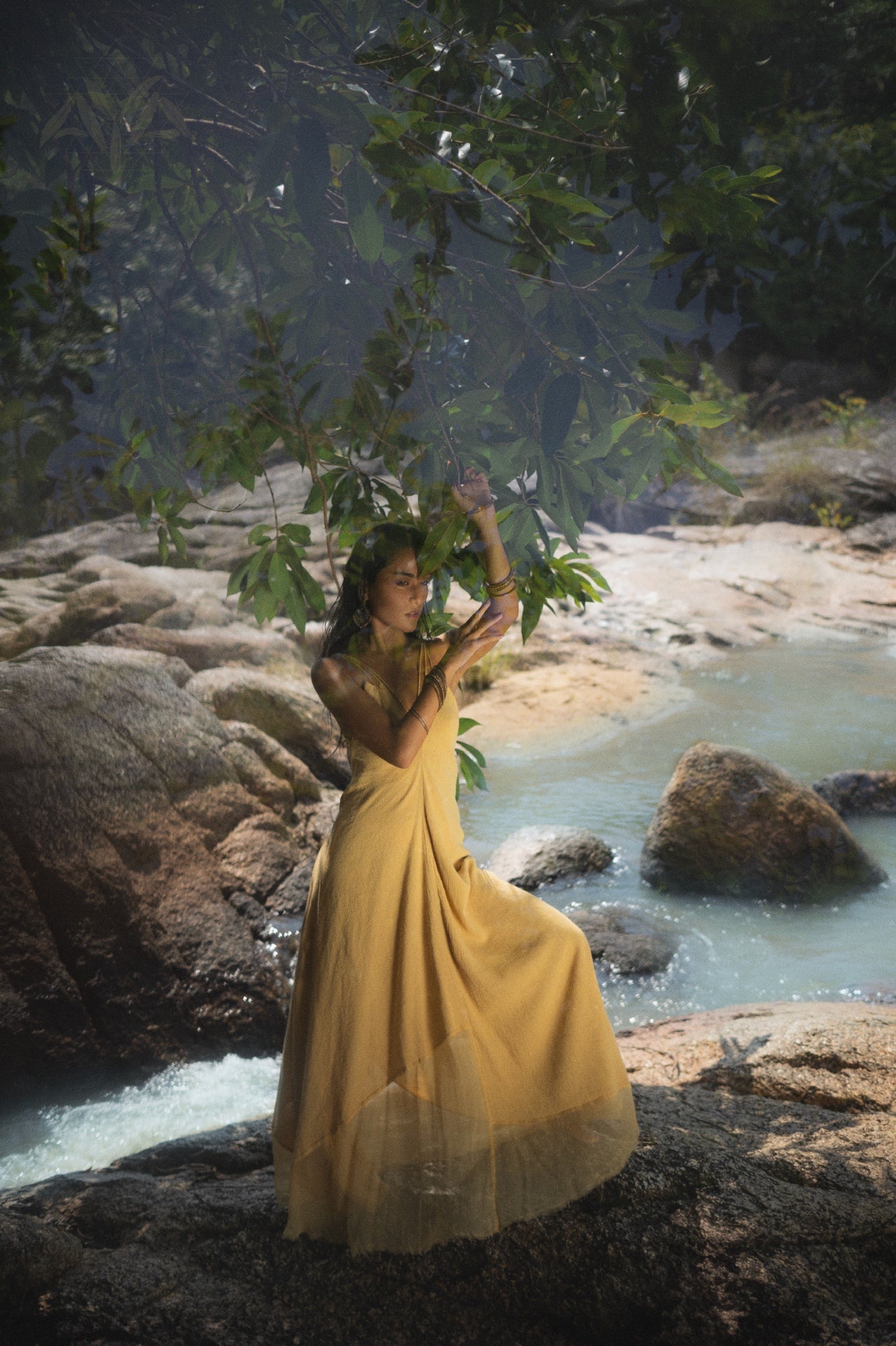 A woman stands on rocks near a serene stream, wearing the Powder Yellow Padma Dress by AYA Sacred Wear. Surrounded by greenery and soft sunlight, she gently holds leafy branches above, creating a tranquil scene in this boho backless, sleeveless elegant dress.