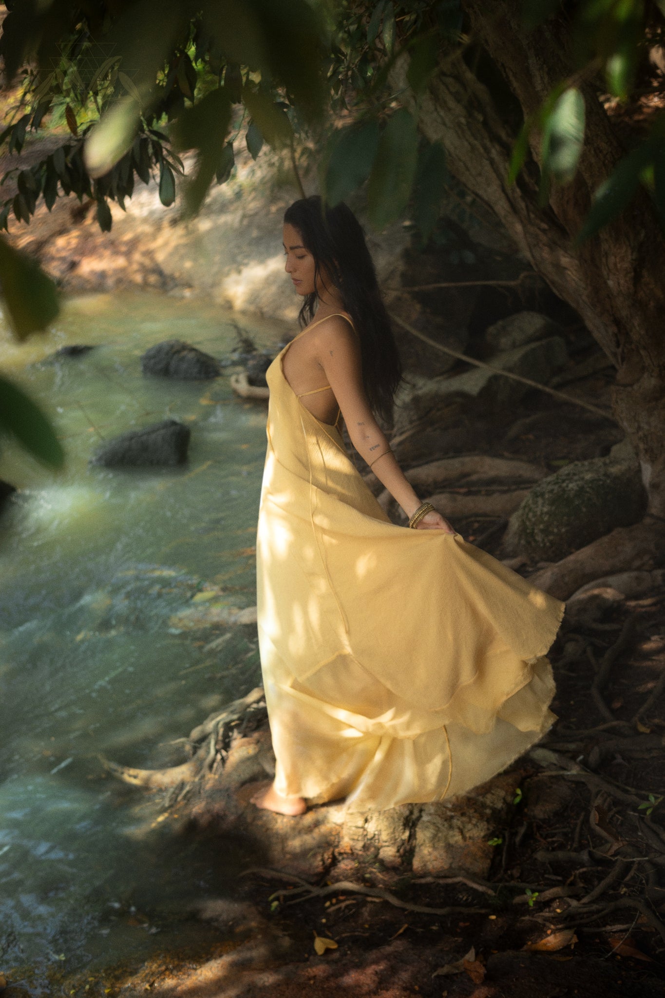 A person in an AYA Sacred Wear Powder Yellow Padma Dress, a boho backless and sleeveless elegant gown, stands by a serene stream. Overhanging trees filter sunlight through the leaves, casting dappled light and creating a natural atmosphere.