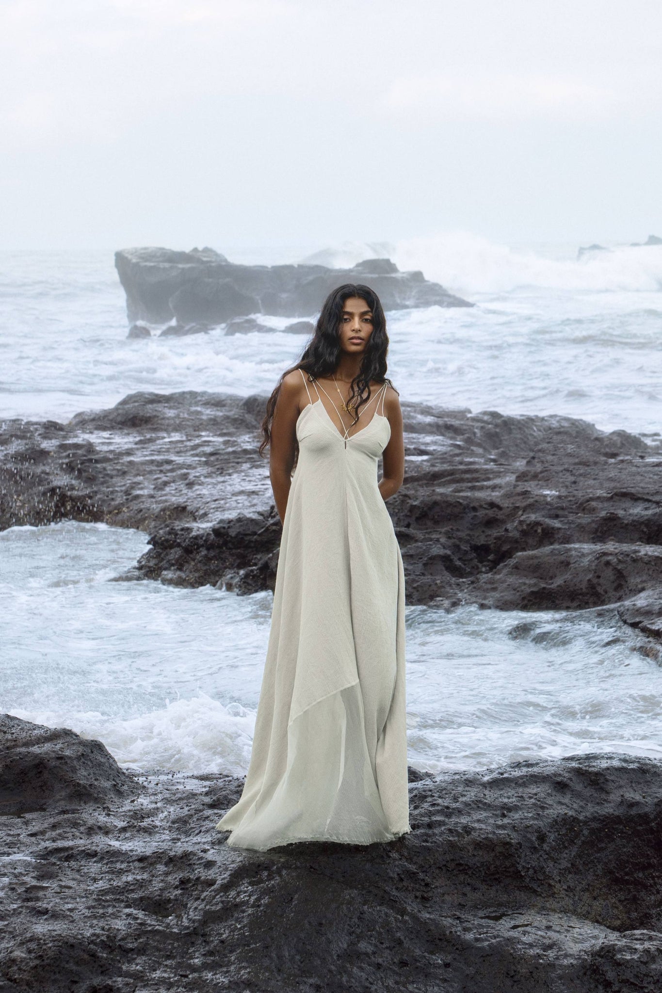 A woman in the Powder Blue Padma Dress by AYA Sacred Wear stands on rocky terrain by the ocean. Waves crash behind her under a cloudy sky, her long dark hair flowing as she gazes serenely into the distance.