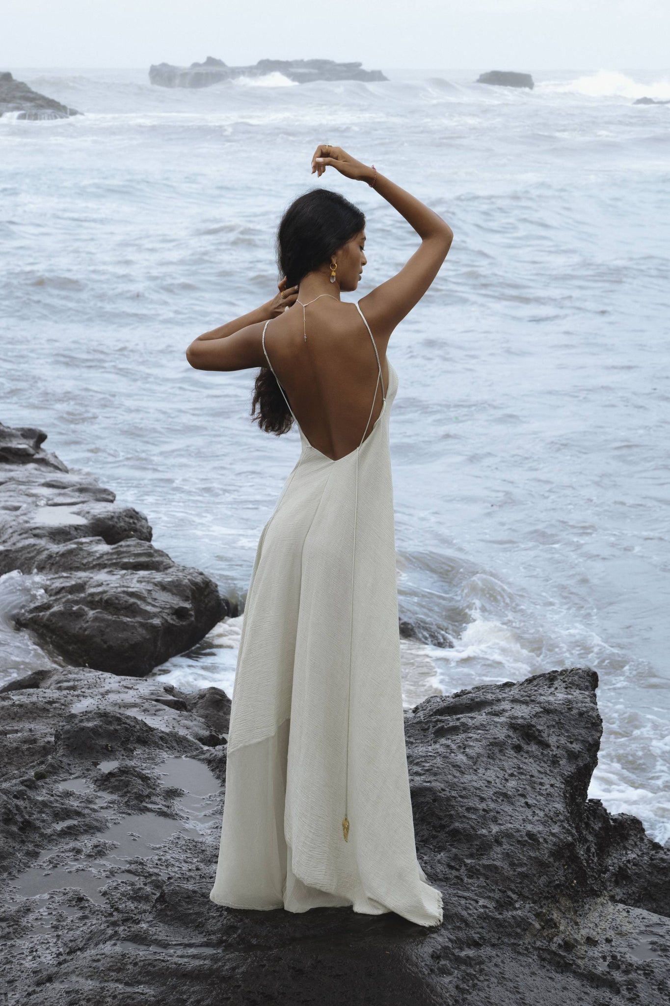 A woman in a Powder Blue Padma Dress from AYA Sacred Wear stands on rocky terrain by the ocean. Her long dark hair falls down her back, arms raised above her head. The slightly rough sea crashes against the rocks under a cloudy sky, creating a mystical scene.