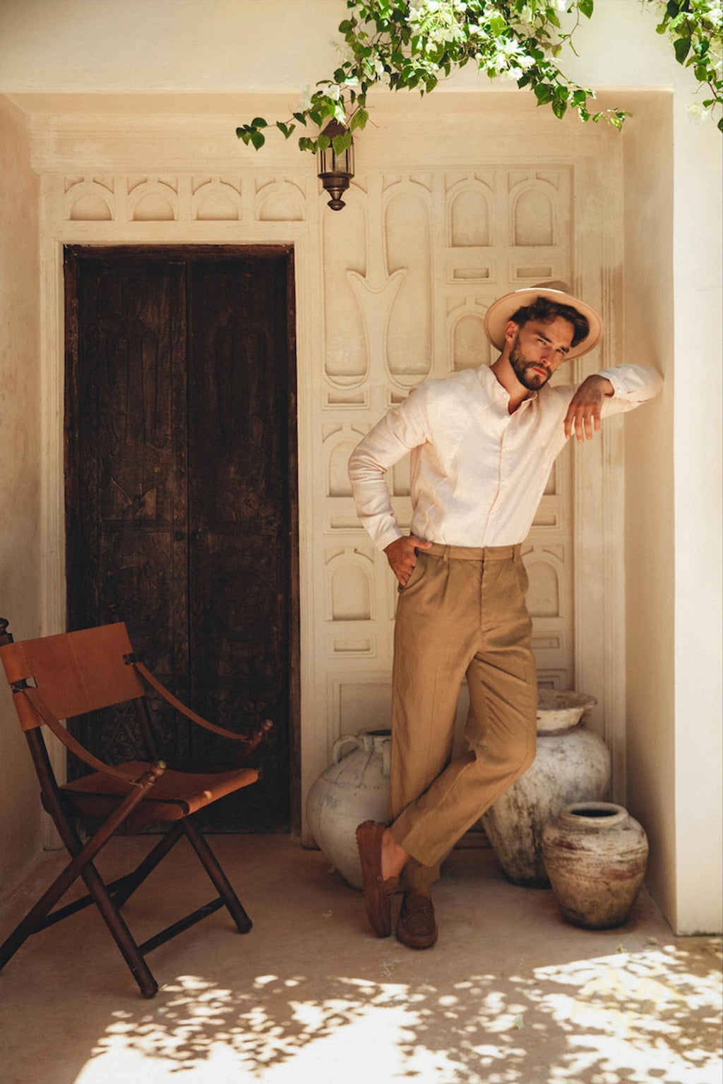 Dressed in a Pale Pink Linen Classic Minimalist Shirt from AYA Sacred Wear and tan pants, a man complements his outfit with a beige hat as he leans against an ornate rustic wall. Large clay pots surround him, while a wooden chair sits to his left, and green leaves rustle softly under the glow of a hanging lantern.