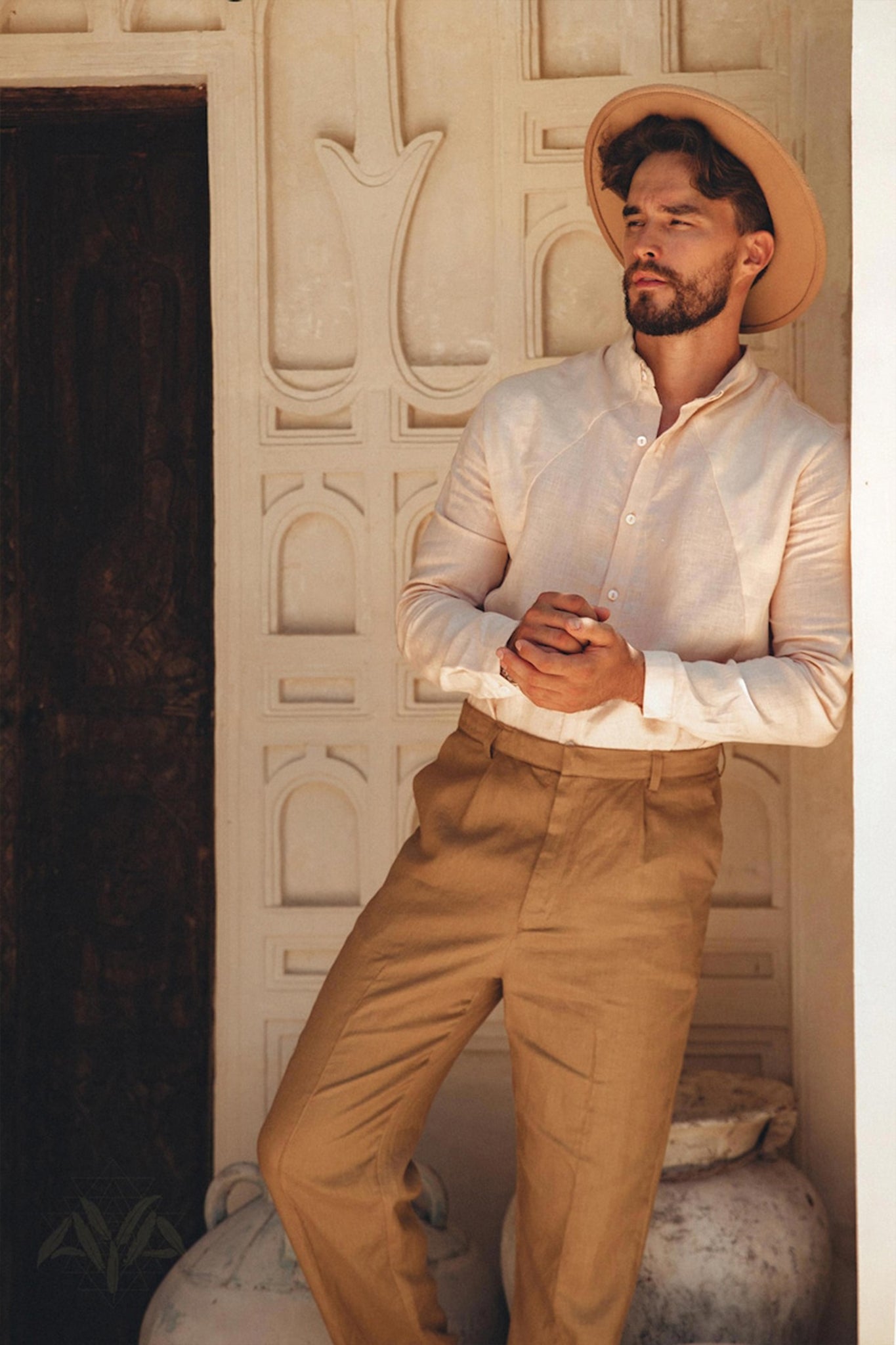 A man in a Pale Pink Linen Classic Minimalist Shirt from AYA Sacred Wear and tan trousers leans against a wall with decorative patterns. He has a contemplative expression beneath his wide-brimmed hat. Large pots adorn the background, complementing the natural charm of his botanically dyed attire.