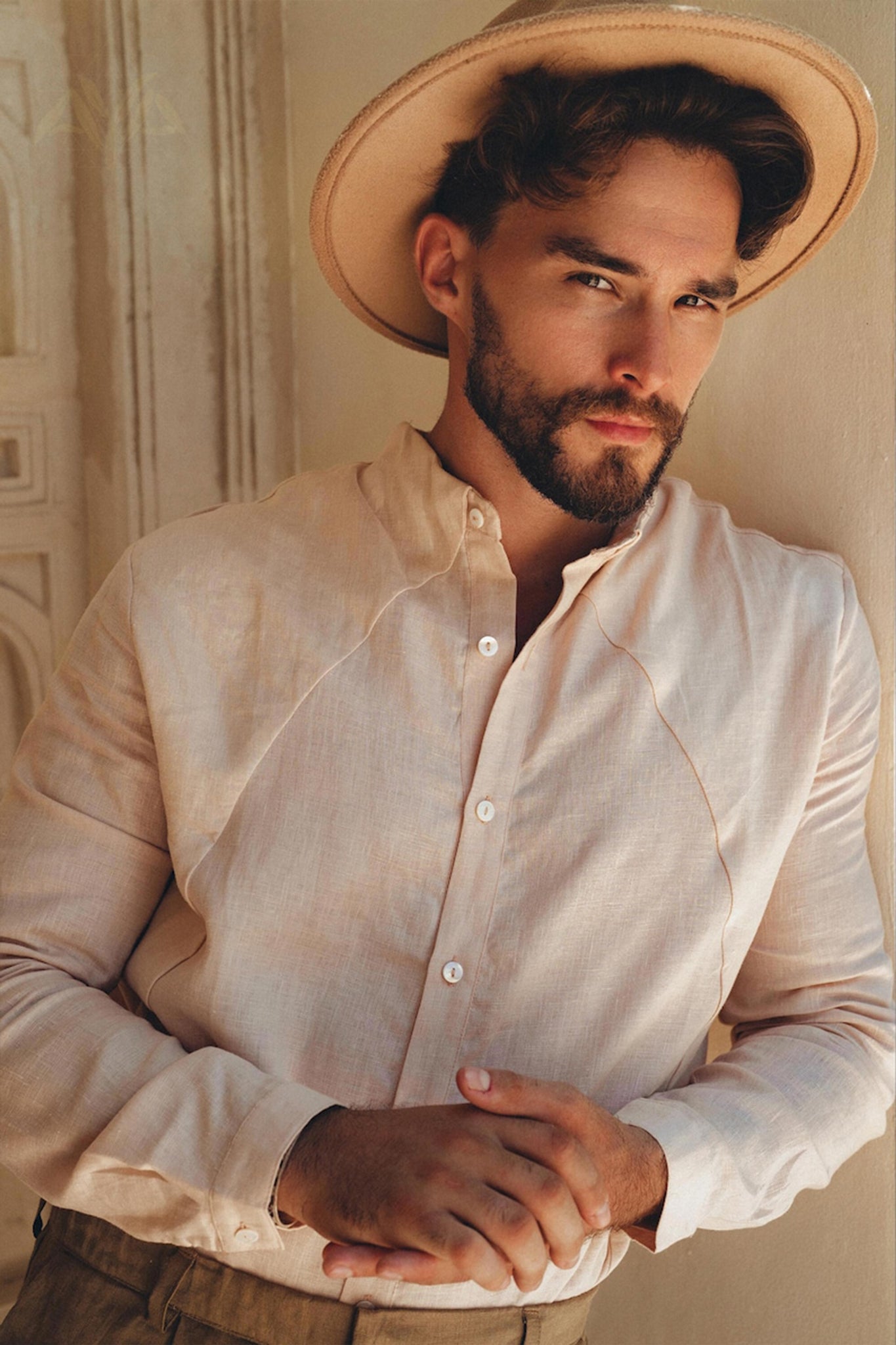 A man with a beard and a neutral expression is wearing the Pale Pink Linen Classic Minimalist Shirt for Men by AYA Sacred Wear, botanically dyed for an organic touch, paired with a wide-brimmed hat. He stands indoors against a light-colored wall, hands clasped in front of him.