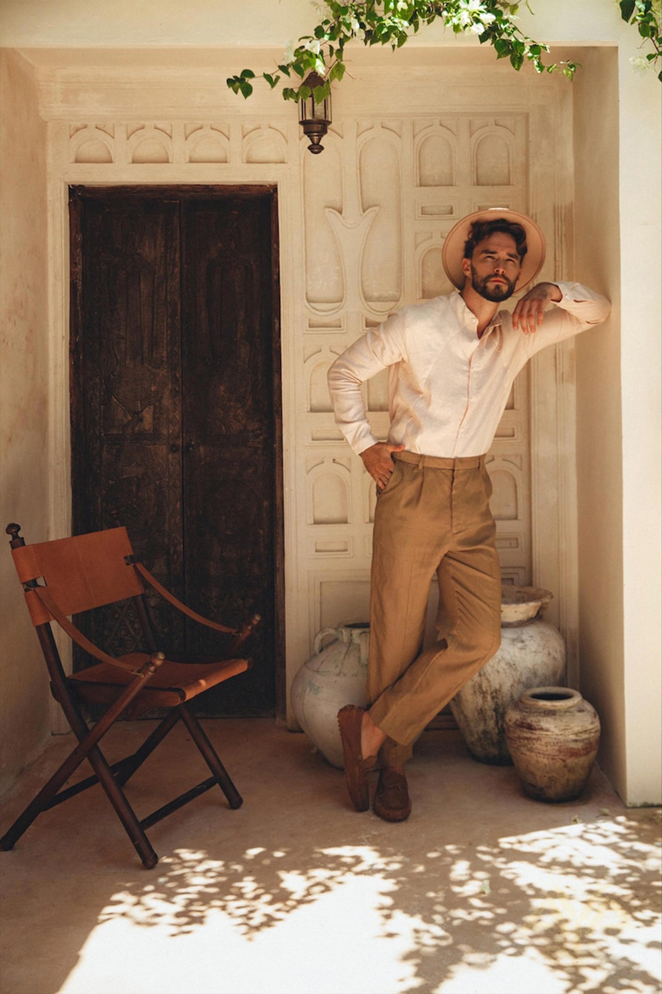 A man in a Pale Pink Linen Classic Minimalist Shirt by AYA Sacred Wear, beige trousers, and a wide-brimmed hat leans against a textured wall in a sunlit alcove. Beside him are large ceramic pots and a wooden chair. A small lantern hangs above, with greenery peeking into the frame.