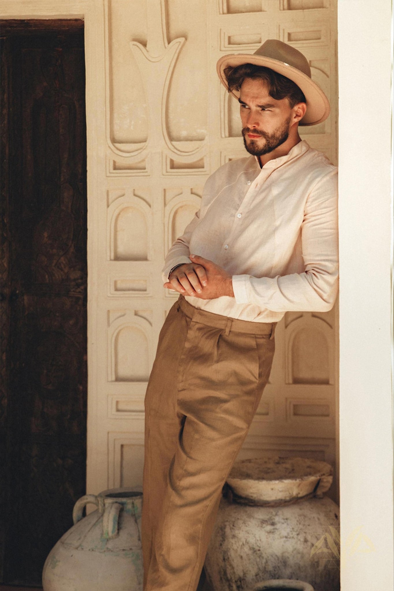 A man with a beard, dressed in a Pale Pink Linen Classic Minimalist Shirt by AYA Sacred Wear and khaki pants, leans against an ornate wall with his hands clasped. A wide-brimmed hat shades his eyes as two large ceramic vases rest on the ground nearby.