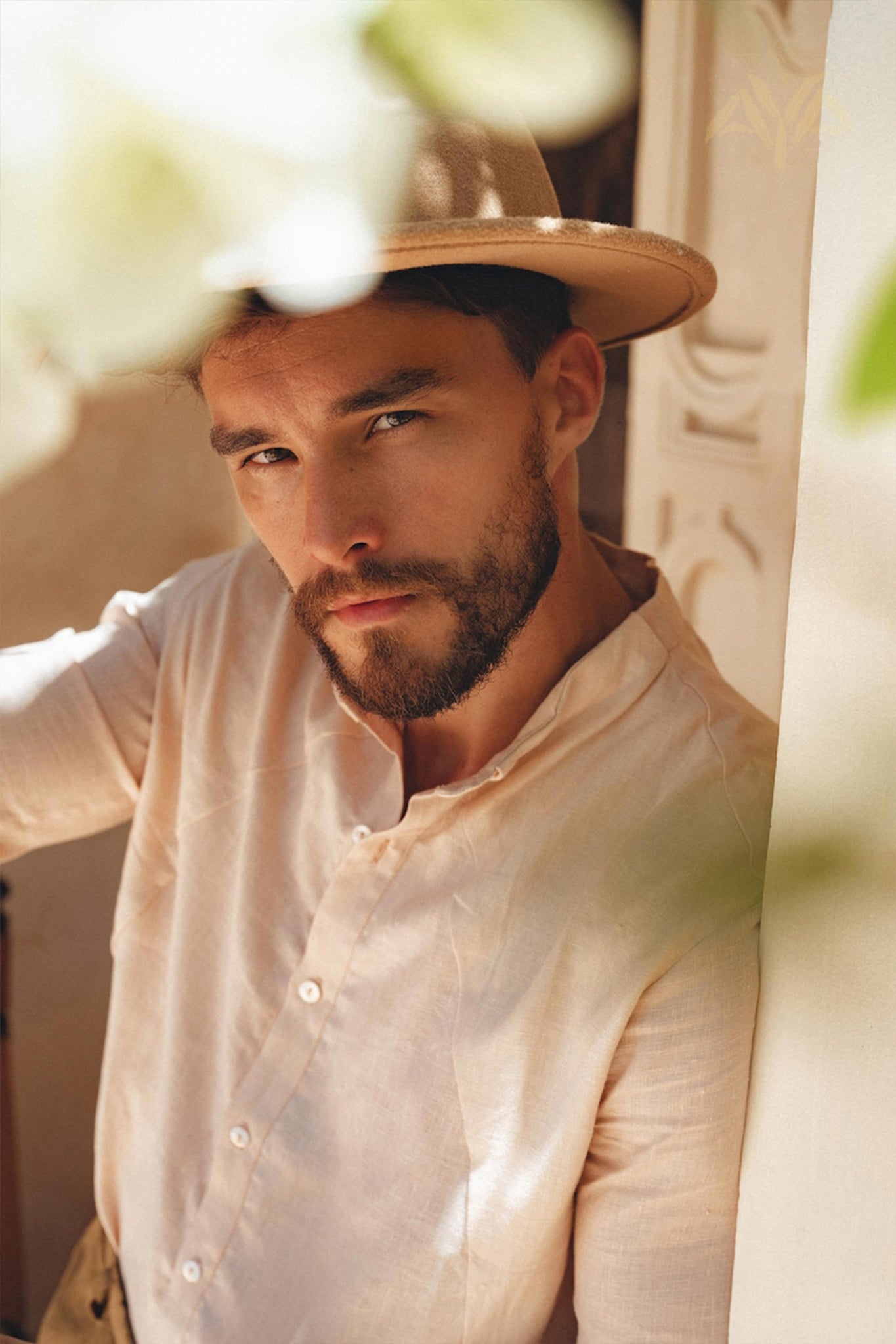 A bearded man wearing an AYA Sacred Wear Pale Pink Linen Classic Minimalist Shirt and a hat gazes intently at the camera. He's leaning against a wall with intricate patterns, enveloped by soft natural light and blurred white flowers in the foreground.