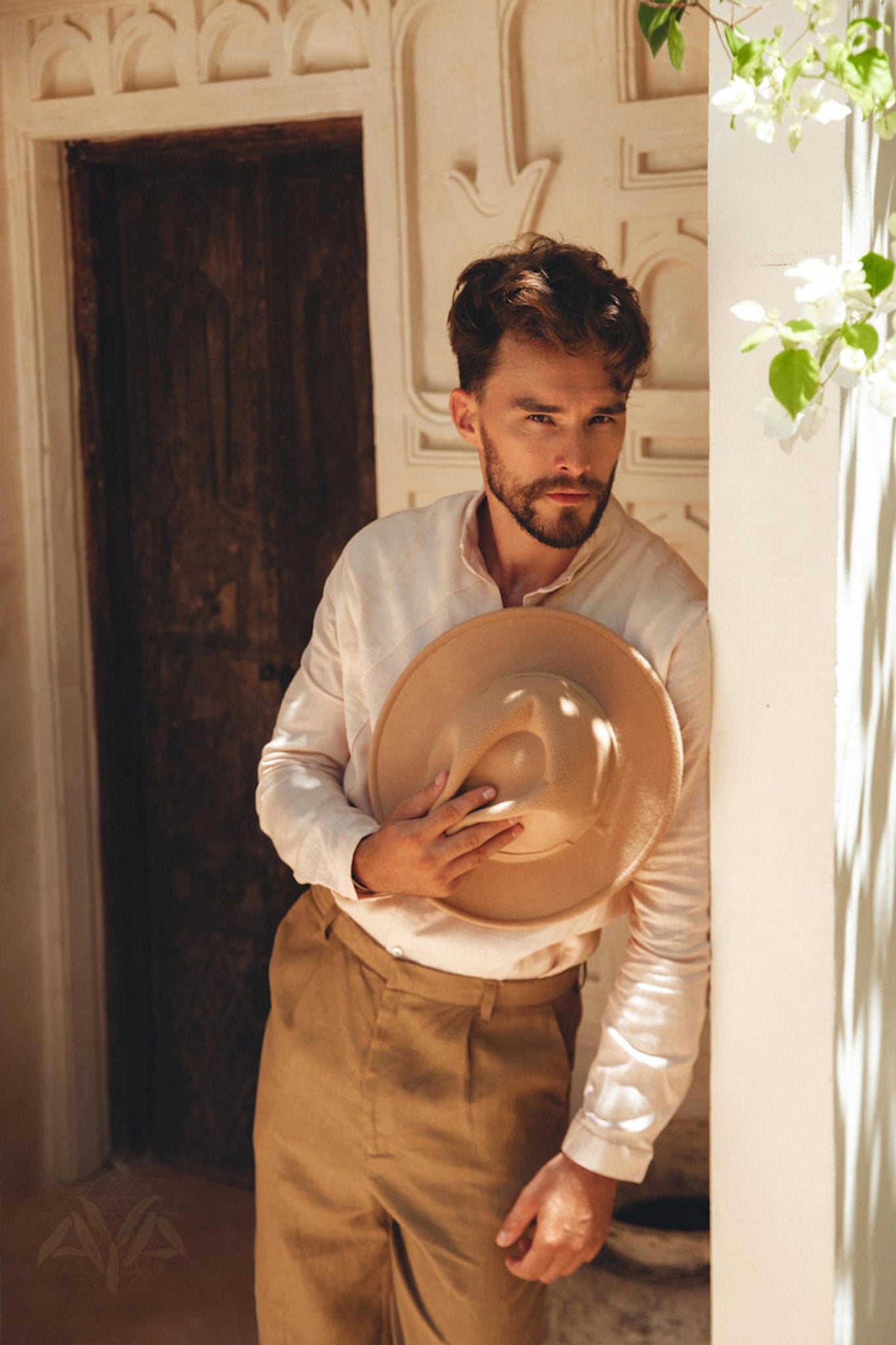 A man with a beard leans against a wall, holding a hat. He's wearing an AYA Sacred Wear Pale Pink Linen Classic Minimalist Shirt and brown pants. The setting is rustic, featuring a wooden door with carved details. A plant with white flowers is visible at the top right.