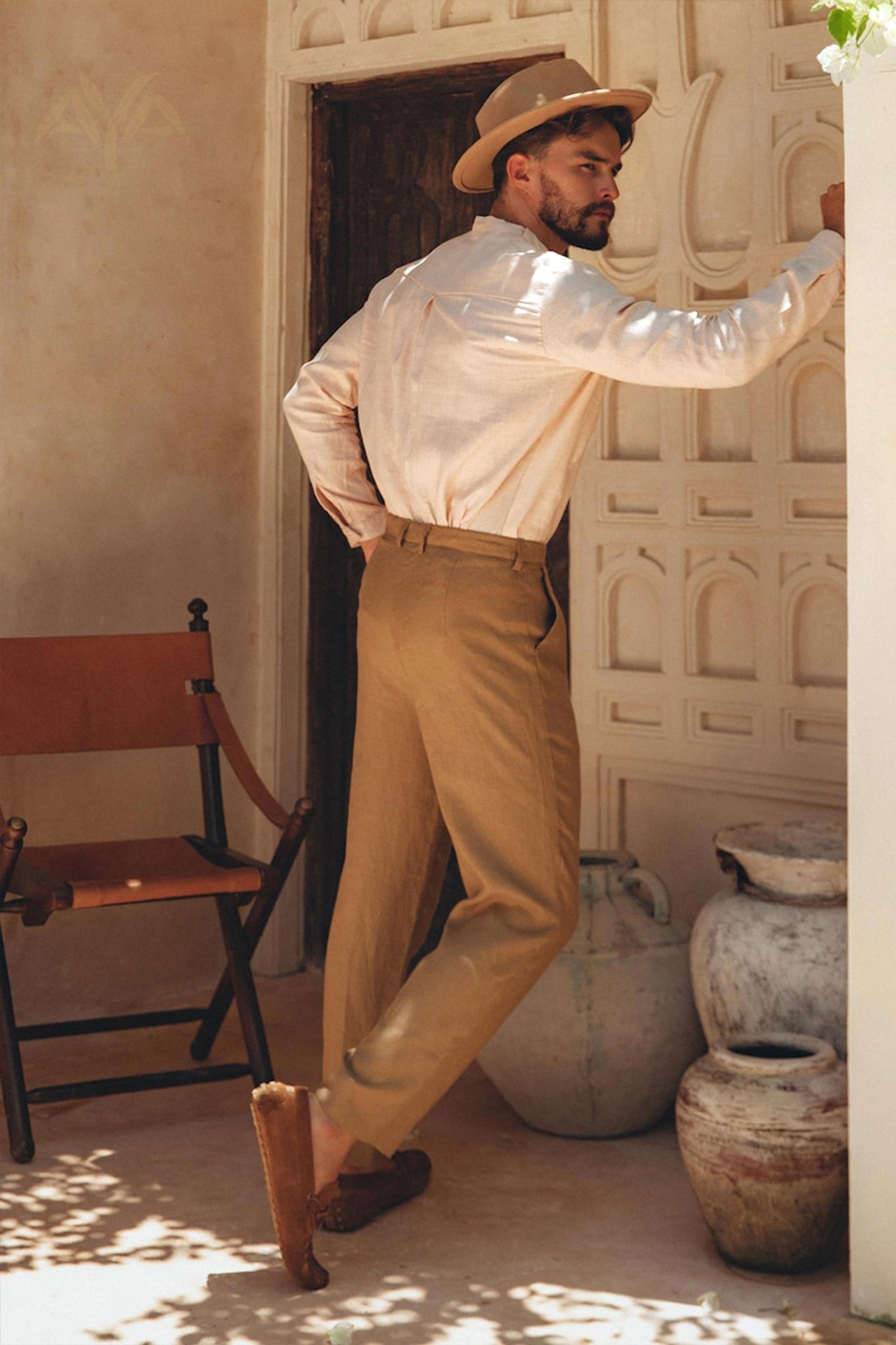 A man with a beard, dressed in a Pale Pink Linen Classic Minimalist Shirt for Men by AYA Sacred Wear, beige pants, and slippers, leans against a textured wall embellished with intricate patterns. A rustic chair and large clay pots are positioned nearby, complementing the earthy aesthetic.