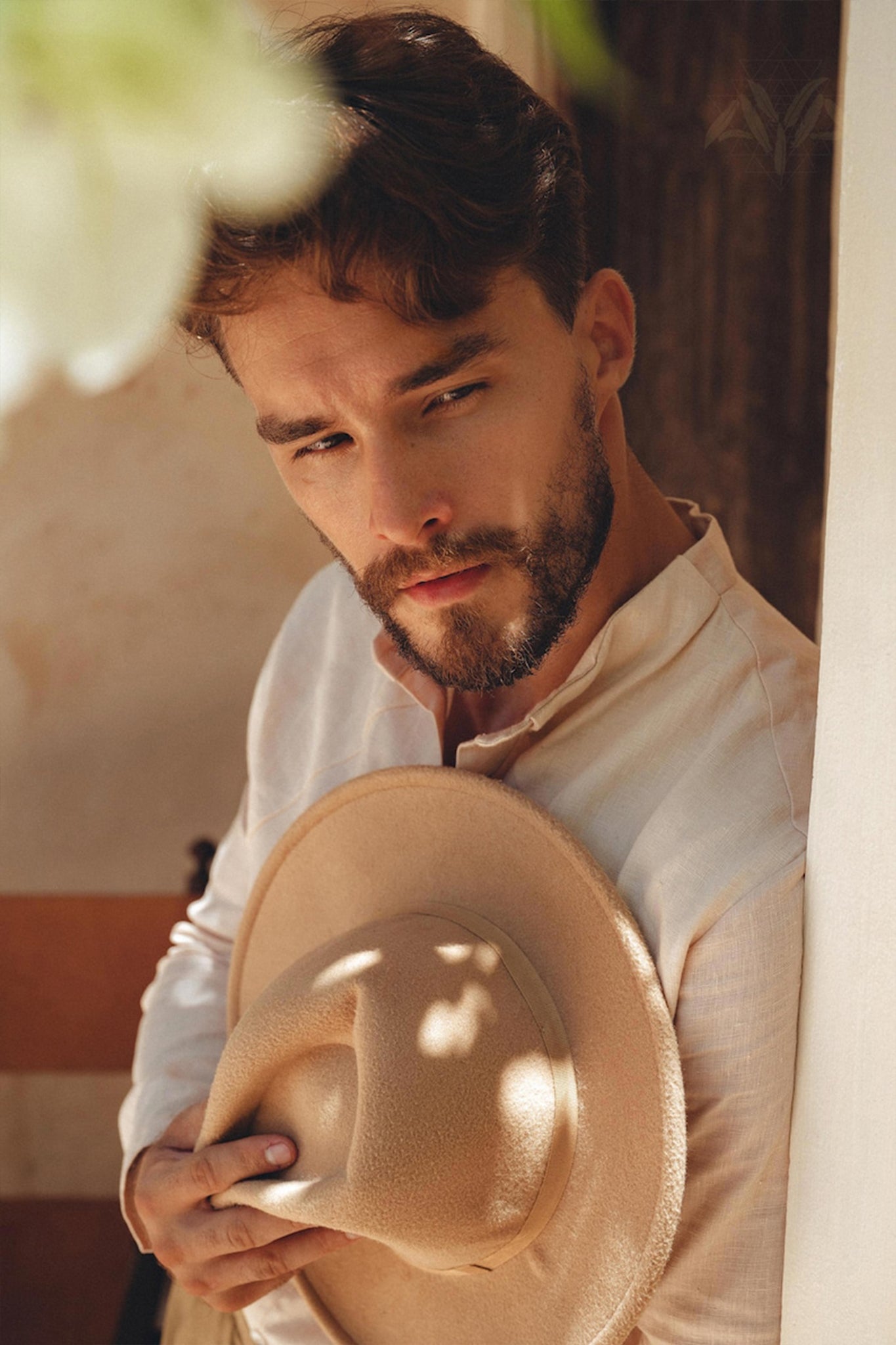 A bearded man wearing an AYA Sacred Wear Pale Pink Linen Classic Minimalist Shirt holds a tan hat against his chest. The shirt, botanically dyed in a soft hue, complements the tranquil setting as he gazes thoughtfully to the side.