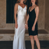 Two women are standing barefoot indoors, holding hands. One is dressed in a flowing bohemian dress in white, while the other wears the sleek Peace Silk Slip Dress by AYA Sacred Wear, crafted from natural silk in a black mini style. They pose before a textured wall with a dark window behind them, both gazing at the camera with serene expressions.