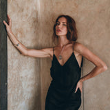 A woman with long hair stands against a textured wall, dressed in the elegant Peace Silk Slip Dress by AYA Sacred Wear. She poses confidently with one hand on her hip, leaning against the wall with her other arm extended and wearing a necklace as she gazes into the distance.