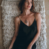 A woman wearing a black Peace Silk Mini Dress by AYA Sacred Wear leans against an intricately woven macrame wall hanging. She accessorizes with a silver necklace and gazes thoughtfully to the side, set against a neutral-toned backdrop.