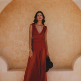 A model stands elegantly in a backless red rose jumpsuit made of silk and cotton, with a deep V-neck and flowing design. She poses against a soft, light-colored background, showcasing the stylish evening overall romper. The outfit flares at the bottom, complemented by a black hat held in her hand, capturing a chic and sophisticated look.