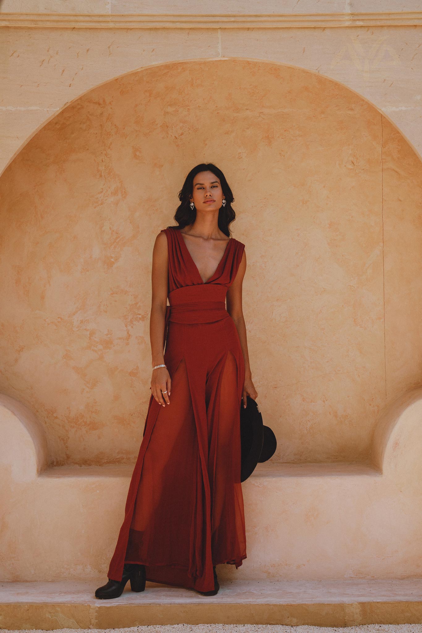 A model stands elegantly in a backless red rose jumpsuit made of silk and cotton, with a deep V-neck and flowing design. She poses against a soft, light-colored background, showcasing the stylish evening overall romper. The outfit flares at the bottom, complemented by a black hat held in her hand, capturing a chic and sophisticated look.
