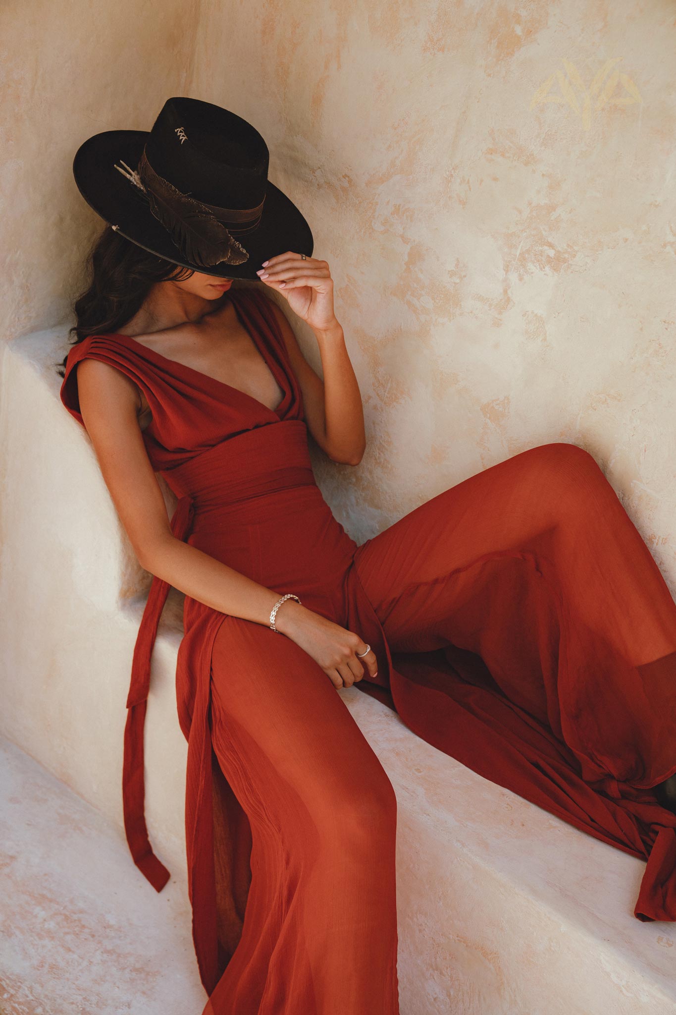 A woman seated against a textured wall in a backless, flowing red rose jumpsuit made from a silk-cotton blend. She is wearing a black hat adorned with feathers, with her hair cascading down one side. The jumpsuit features a deep V-neckline and tied waist, highlighting an elegant silhouette. The overall look is chic and stylish, perfect for an evening event.