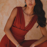 A model poses wearing a backless evening jumpsuit in a rich red rose color made from silk and cotton. The garment features deep V-necklines and elegant draping, complemented by chic silver jewelry. The background has a warm, textured finish.