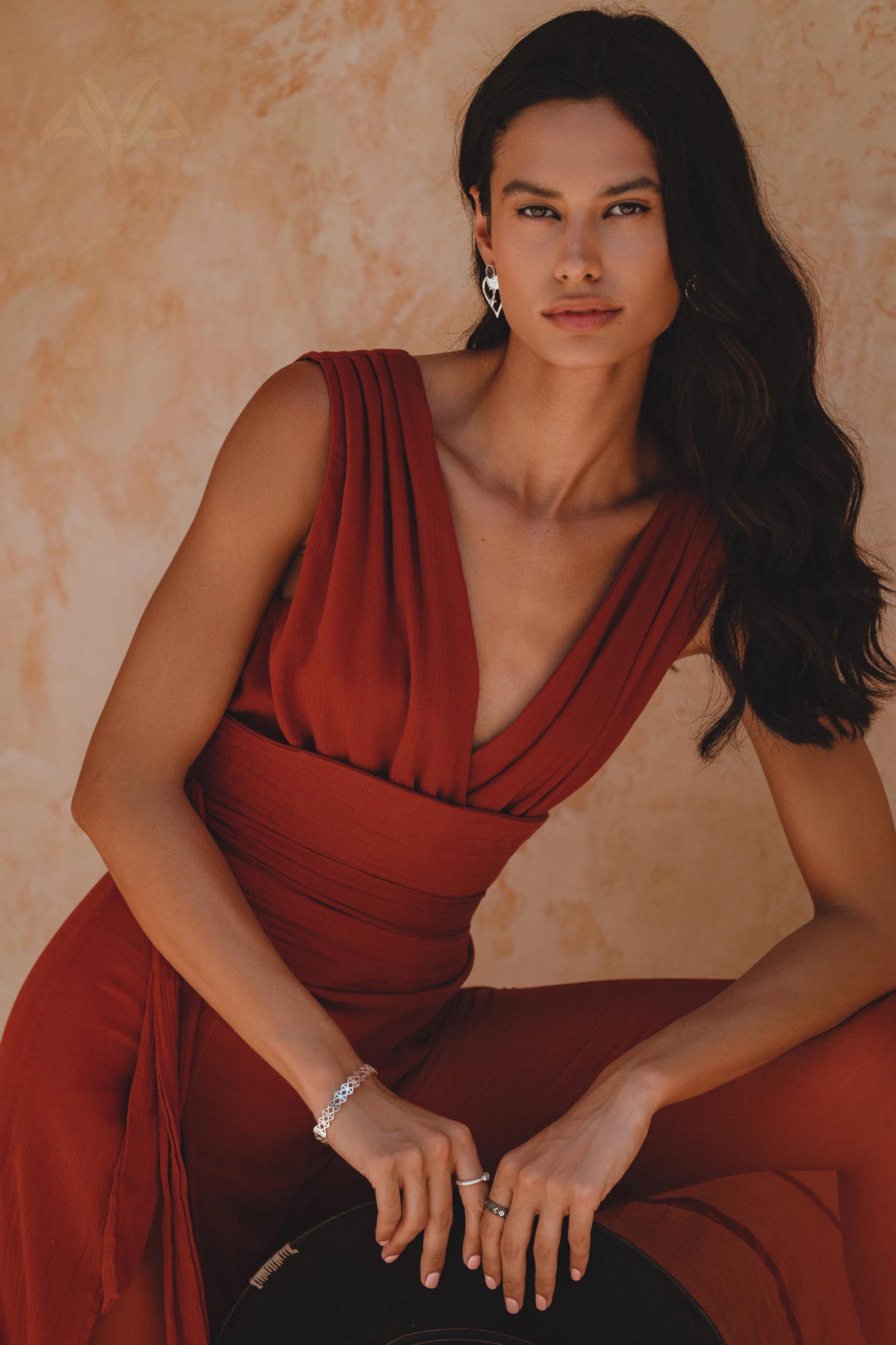 A model poses wearing a backless evening jumpsuit in a rich red rose color made from silk and cotton. The garment features deep V-necklines and elegant draping, complemented by chic silver jewelry. The background has a warm, textured finish.