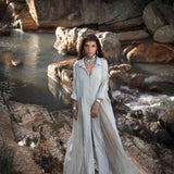 A woman stands on rocks by a stream in a flowing, powder blue Kannika Shirt Dress by AYA Sacred Wear. Multiple necklaces and bracelets adorn her as dappled sunlight filters through trees, highlighting the water and large rocks.