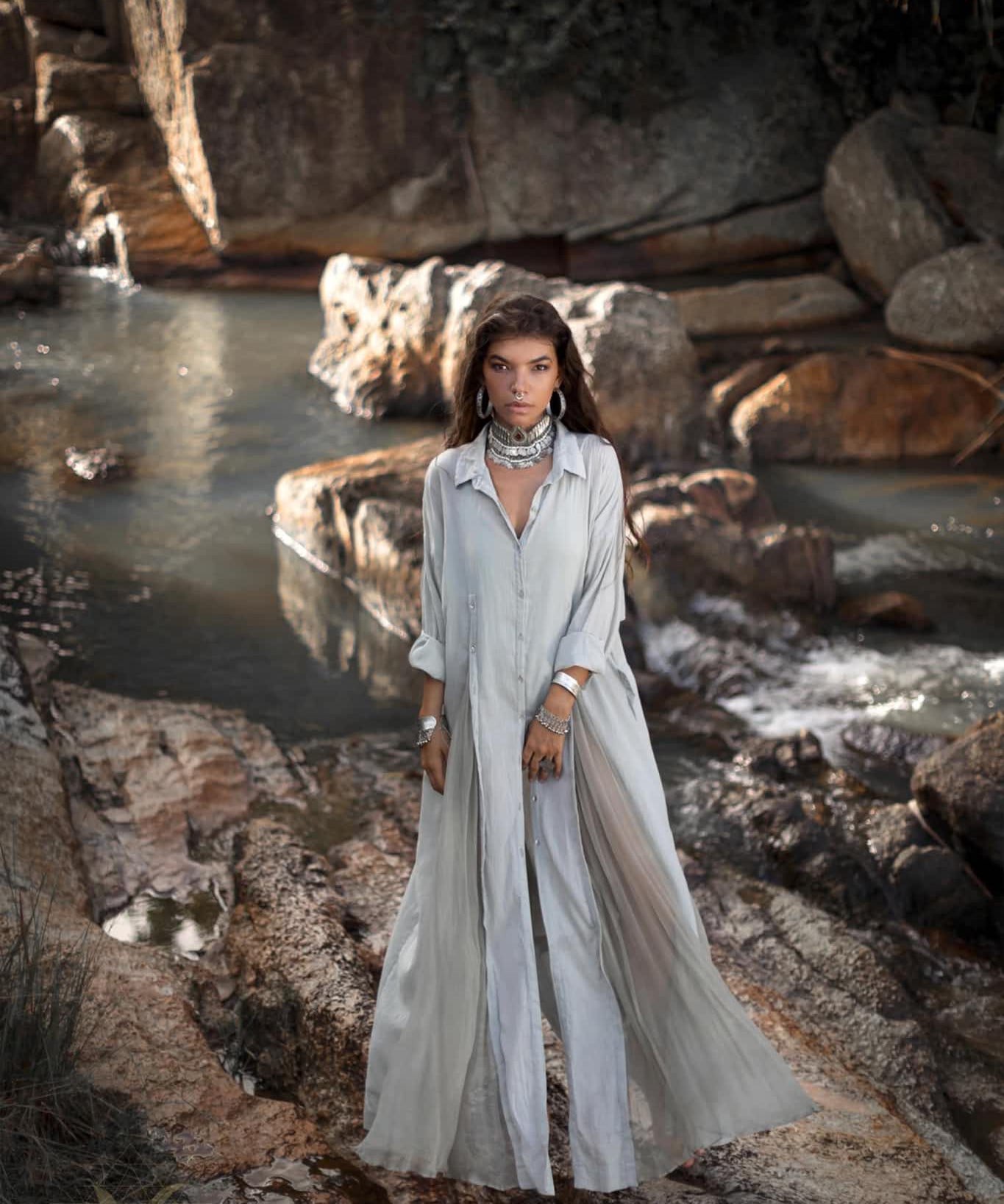 A woman stands on rocks by a stream in a flowing, powder blue Kannika Shirt Dress by AYA Sacred Wear. Multiple necklaces and bracelets adorn her as dappled sunlight filters through trees, highlighting the water and large rocks.