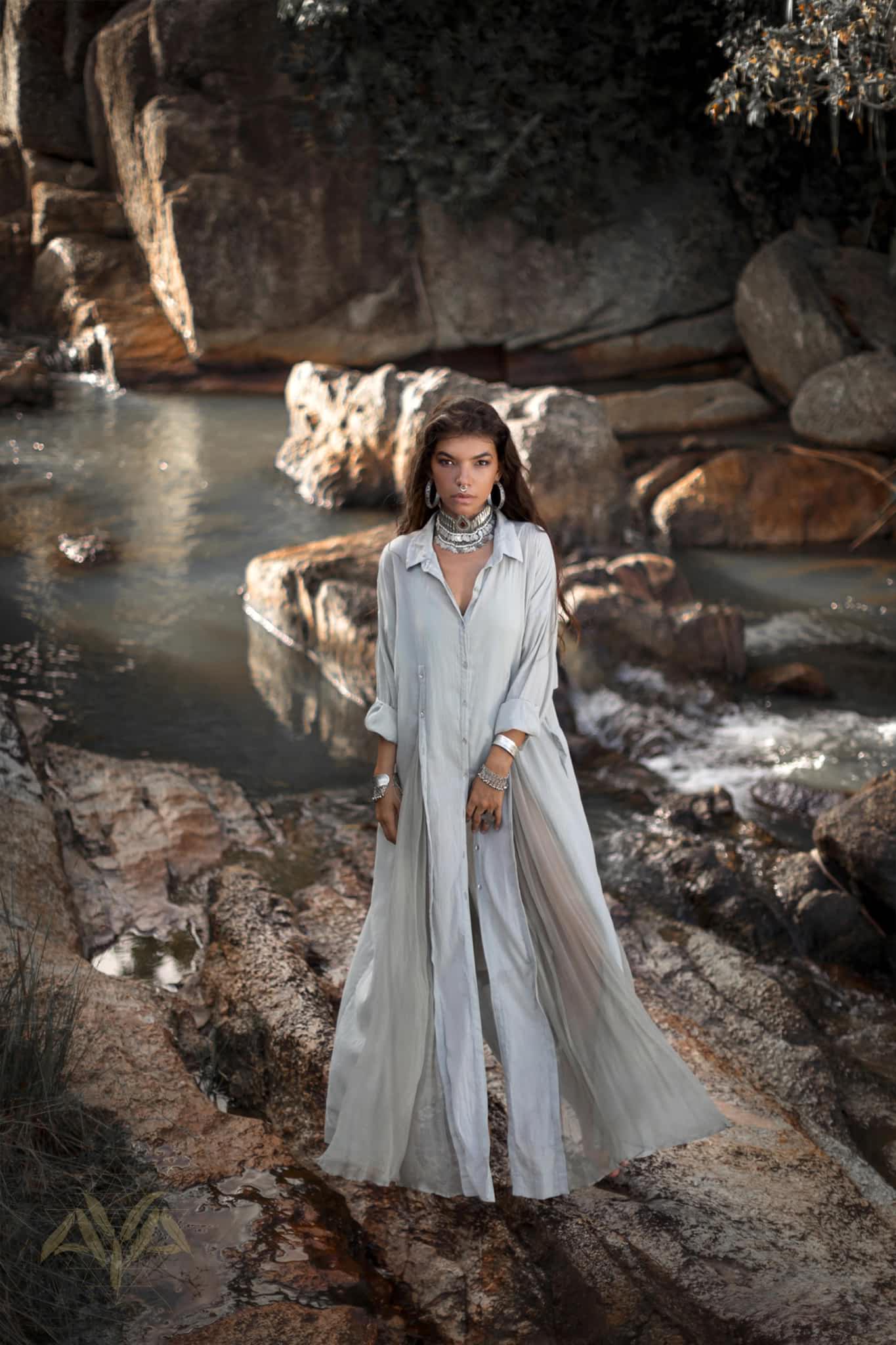 A woman stands on rocks by a stream in a flowing, powder blue Kannika Shirt Dress by AYA Sacred Wear. Multiple necklaces and bracelets adorn her as dappled sunlight filters through trees, highlighting the water and large rocks.