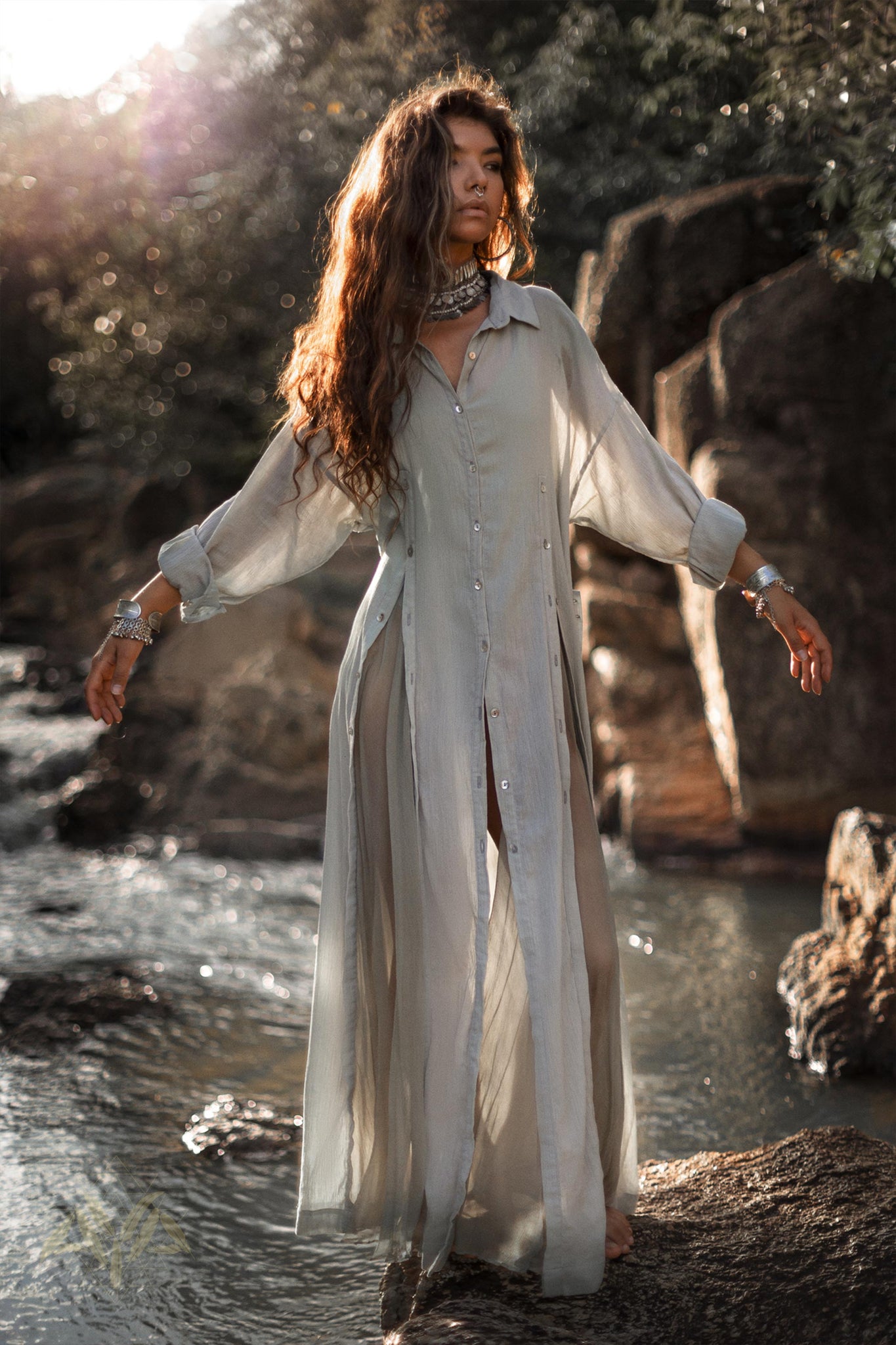 A person with long wavy hair stands on rocks in a flowing, semi-transparent Powder Blue Kannika Shirt Dress by AYA Sacred Wear. They are by a gently flowing stream and surrounded by green foliage. The sunlight filters through the trees, casting a warm glow on the scene.