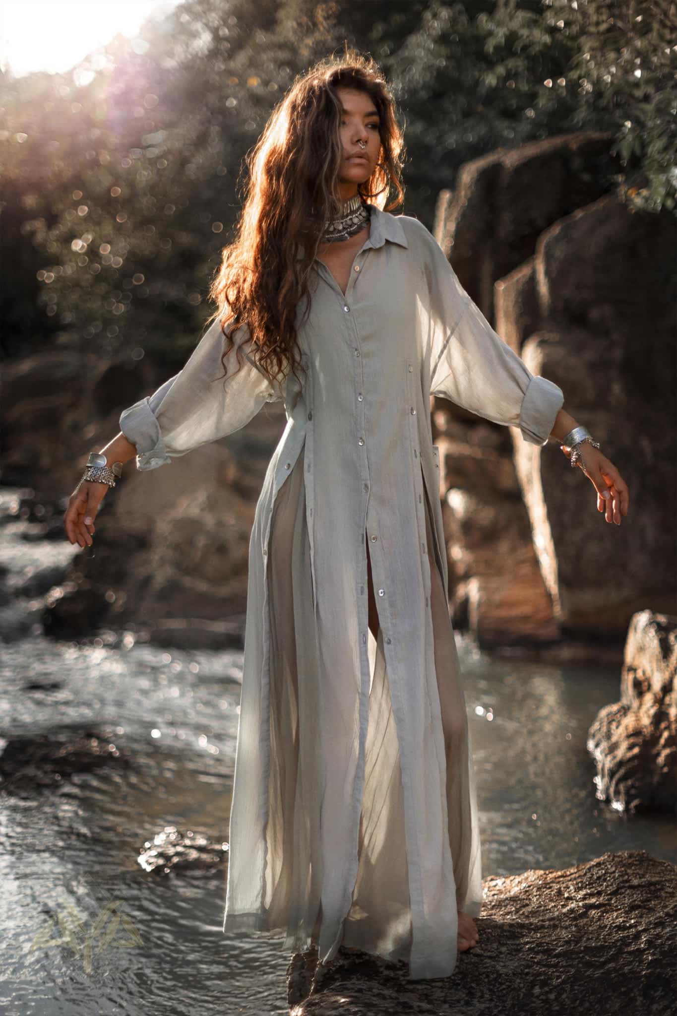 A woman with long hair stands gracefully by a river, wearing an AYA Sacred Wear Powder Blue Kannika Shirt Dress in silk chiffon. Surrounded by rocks and greenery, sunlight streams through the trees, creating a serene and ethereal atmosphere.