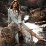 A woman elegantly sits on rocks by a stream in a Powder Blue Kannika Shirt Dress by AYA Sacred Wear. The dress, with its sheer silk chiffon design, flows gracefully as sunlight filters through the trees. She accessorizes with large hoop earrings and a statement necklace, creating a calm, natural scene.