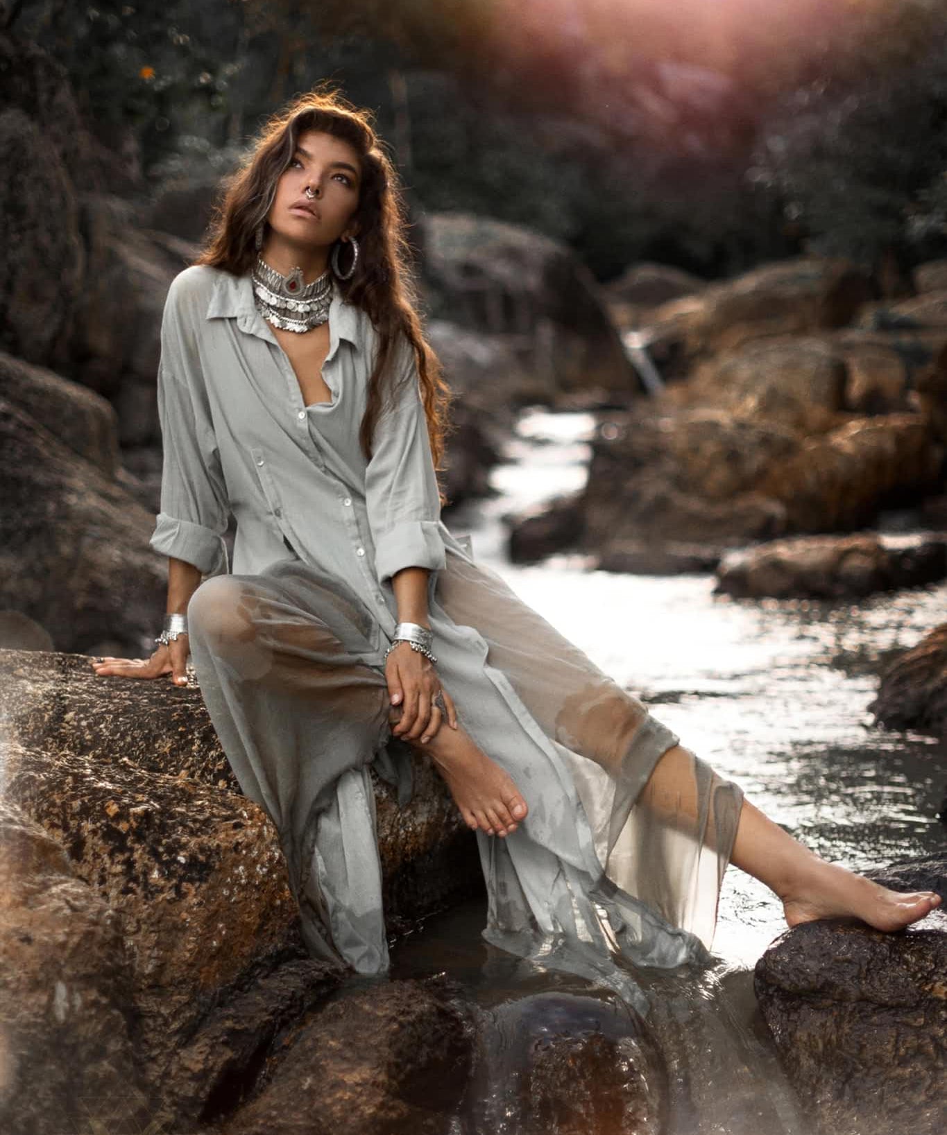 A woman elegantly sits on rocks by a stream in a Powder Blue Kannika Shirt Dress by AYA Sacred Wear. The dress, with its sheer silk chiffon design, flows gracefully as sunlight filters through the trees. She accessorizes with large hoop earrings and a statement necklace, creating a calm, natural scene.