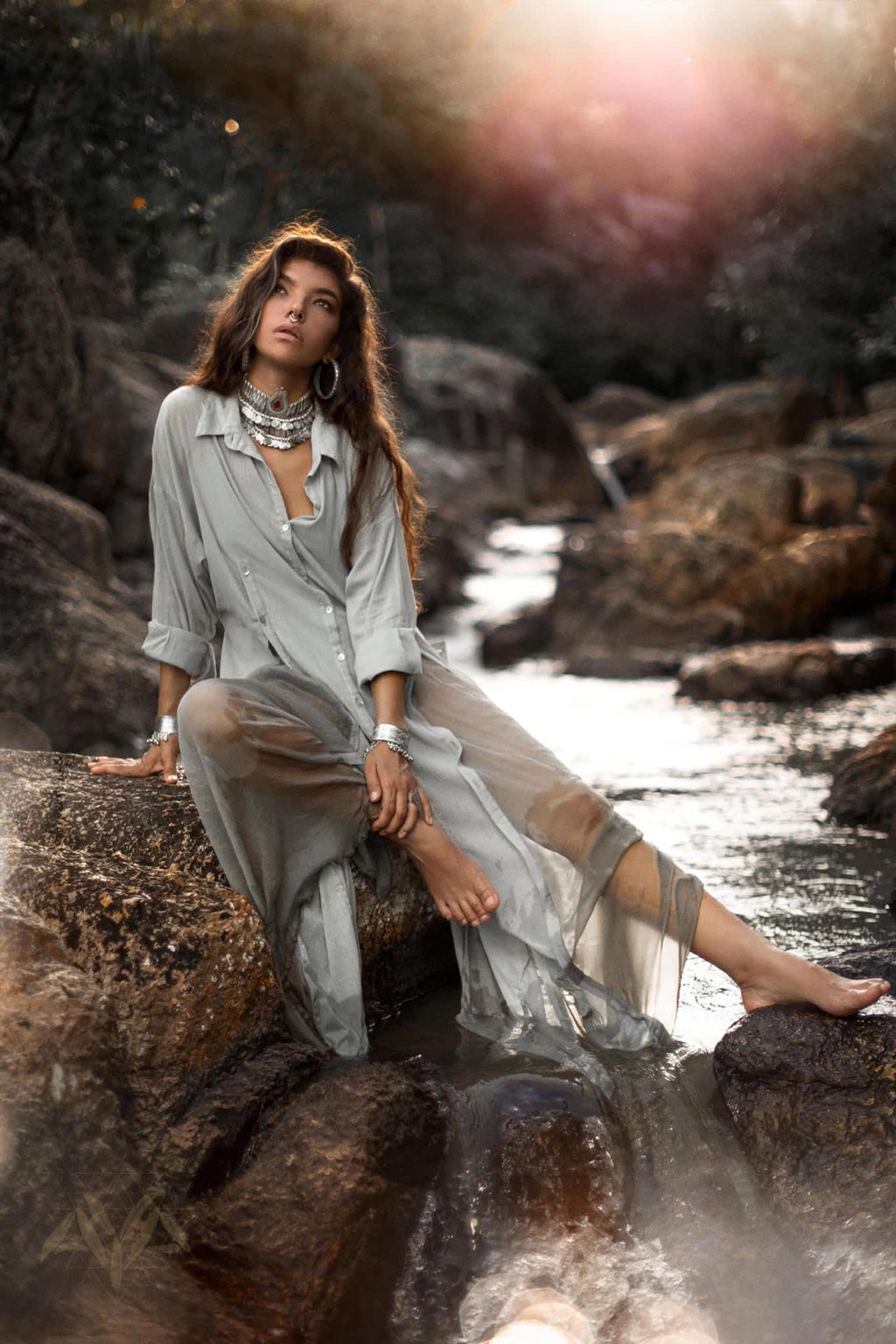 A woman elegantly sits on rocks by a stream in a Powder Blue Kannika Shirt Dress by AYA Sacred Wear. The dress, with its sheer silk chiffon design, flows gracefully as sunlight filters through the trees. She accessorizes with large hoop earrings and a statement necklace, creating a calm, natural scene.