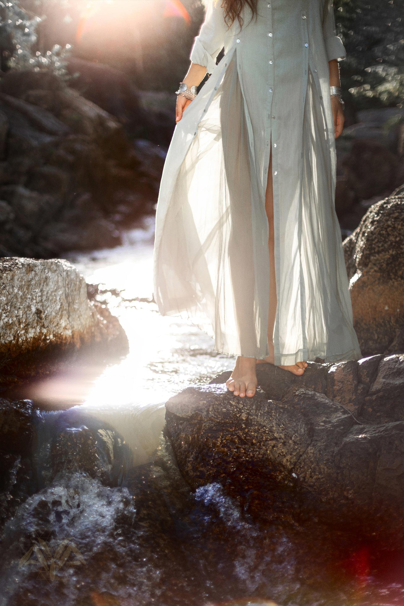 A person stands barefoot on rocks at the edge of a flowing stream. They are wearing an AYA Sacred Wear Powder Blue Kannika Shirt Dress, crafted from organic crinkle cotton that flows in the light breeze. Sunlight filters through the trees, creating a serene and ethereal atmosphere around them, enhanced by botanical dyes.