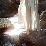 In a serene, nature-inspired scene, someone wearing the Powder Blue Kannika Shirt Dress by AYA Sacred Wear walks barefoot on wet rocks near a stream. Sunlight filters through the trees, casting a warm glow and creating lens flares around the versatile multiway button-down wrap dress.