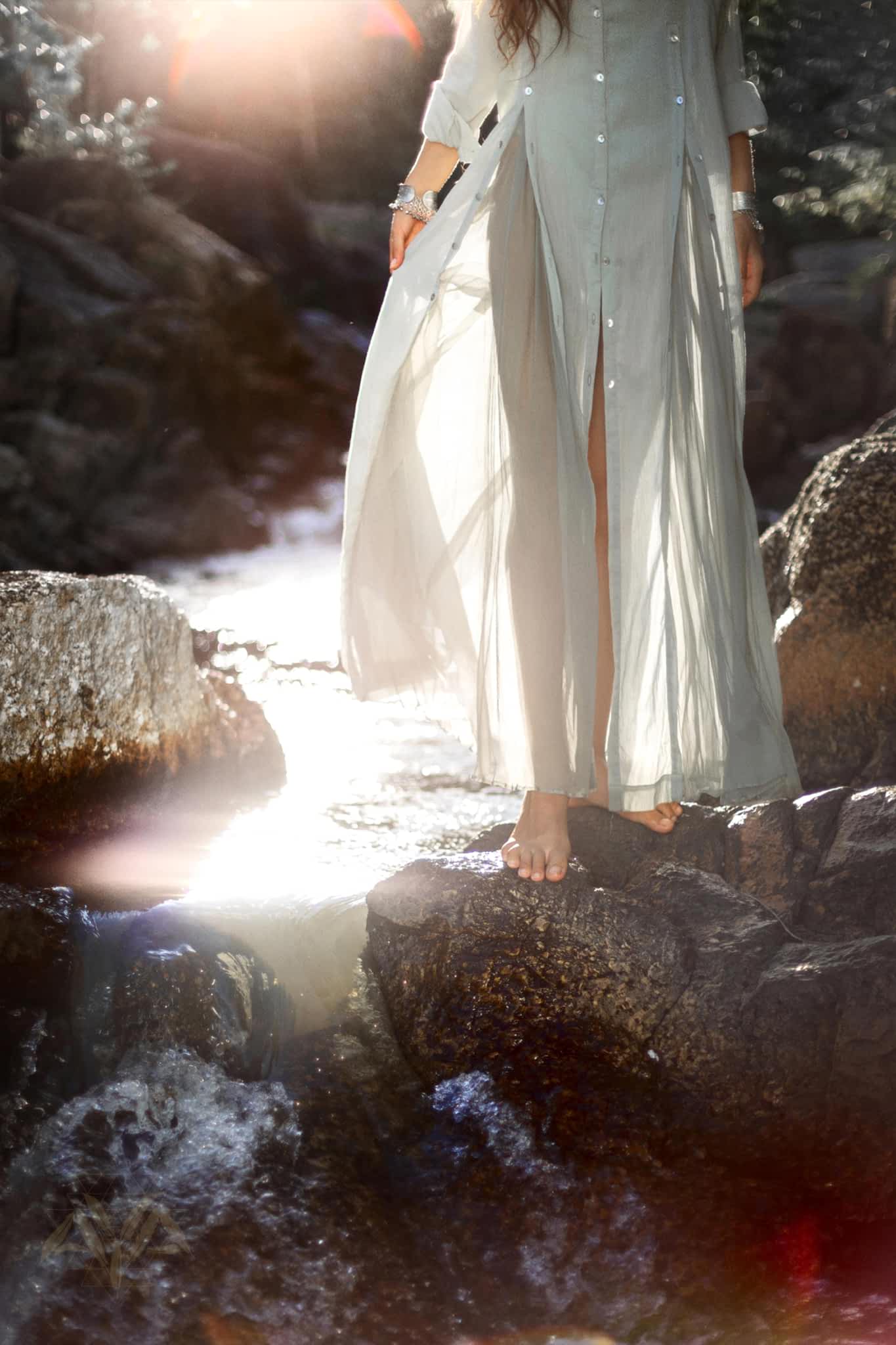 In a serene, nature-inspired scene, someone wearing the Powder Blue Kannika Shirt Dress by AYA Sacred Wear walks barefoot on wet rocks near a stream. Sunlight filters through the trees, casting a warm glow and creating lens flares around the versatile multiway button-down wrap dress.