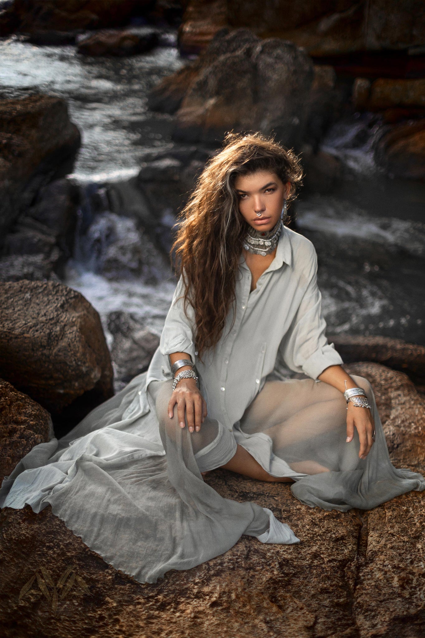 A person with long, wavy hair sits on rocks near water, adorned with multiple pieces of jewelry including necklaces, bracelets, and rings. They are wearing a flowing Powder Blue Kannika Shirt Dress from AYA Sacred Wear, made from organic crinkle cotton and botanical dyes. This versatile dress can be styled as a multiway button down or a wrap cover up. A small waterfall and rugged natural landscape frame their serene presence.