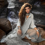A person with long, wavy hair sits on rocks by flowing water, wearing an AYA Sacred Wear Powder Blue Kannika Shirt Dress. Multiple silver bracelets and necklaces enhance their look. The tranquil setting and light casting shadows highlight the dress's silhouette.