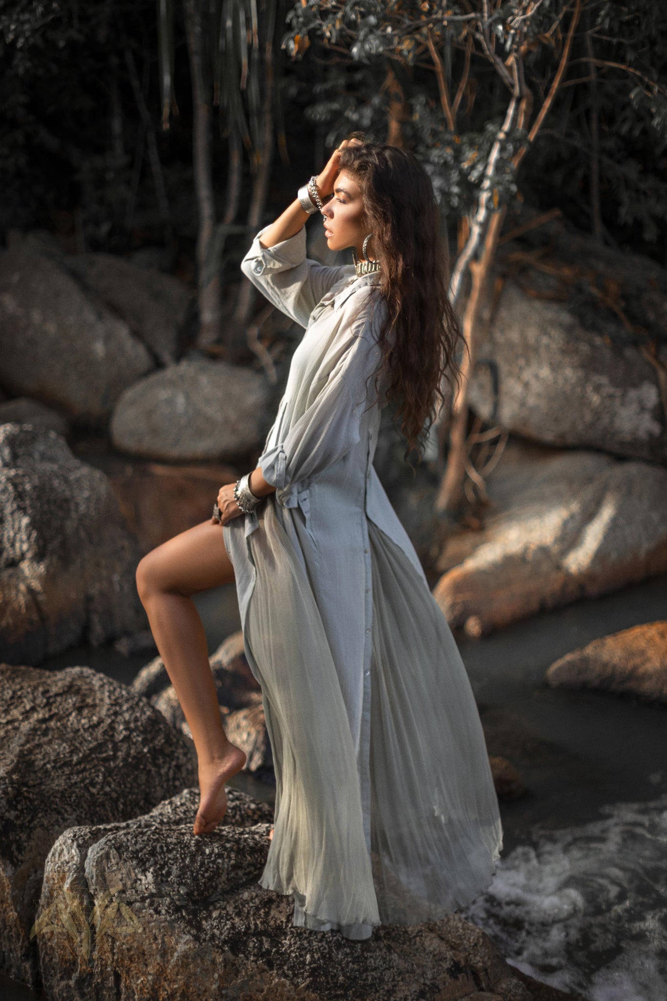 A woman with long, wavy hair stands barefoot on a rock by a river, wearing an AYA Sacred Wear Powder Blue Kannika Shirt Dress. With one arm raised to her forehead, she blends seamlessly into the natural, serene setting of rocks and greenery.