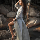 A woman stands barefoot on a rock by a stream, wearing AYA Sacred Wear's Powder Blue Kannika Shirt Dress. Crafted from pure silk chiffon, the multiway button-down dress flutters gently as she gazes into the distance, surrounded by large rocks and lush vegetation.