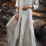 A person stands on rocks in an AYA Sacred Wear Powder Blue Kannika Shirt Dress, a versatile button-down and wrap cover-up made from organic crinkle cotton. The flowing dress billows around them, while bracelets and a necklace adorn their wrists and neck.