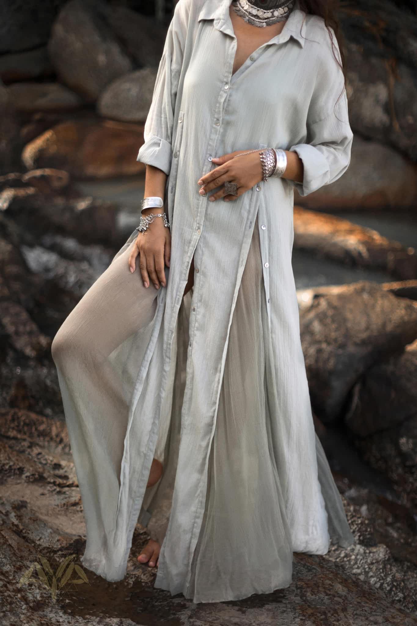 A person stands on rocks in an AYA Sacred Wear Powder Blue Kannika Shirt Dress, a versatile button-down and wrap cover-up made from organic crinkle cotton. The flowing dress billows around them, while bracelets and a necklace adorn their wrists and neck.