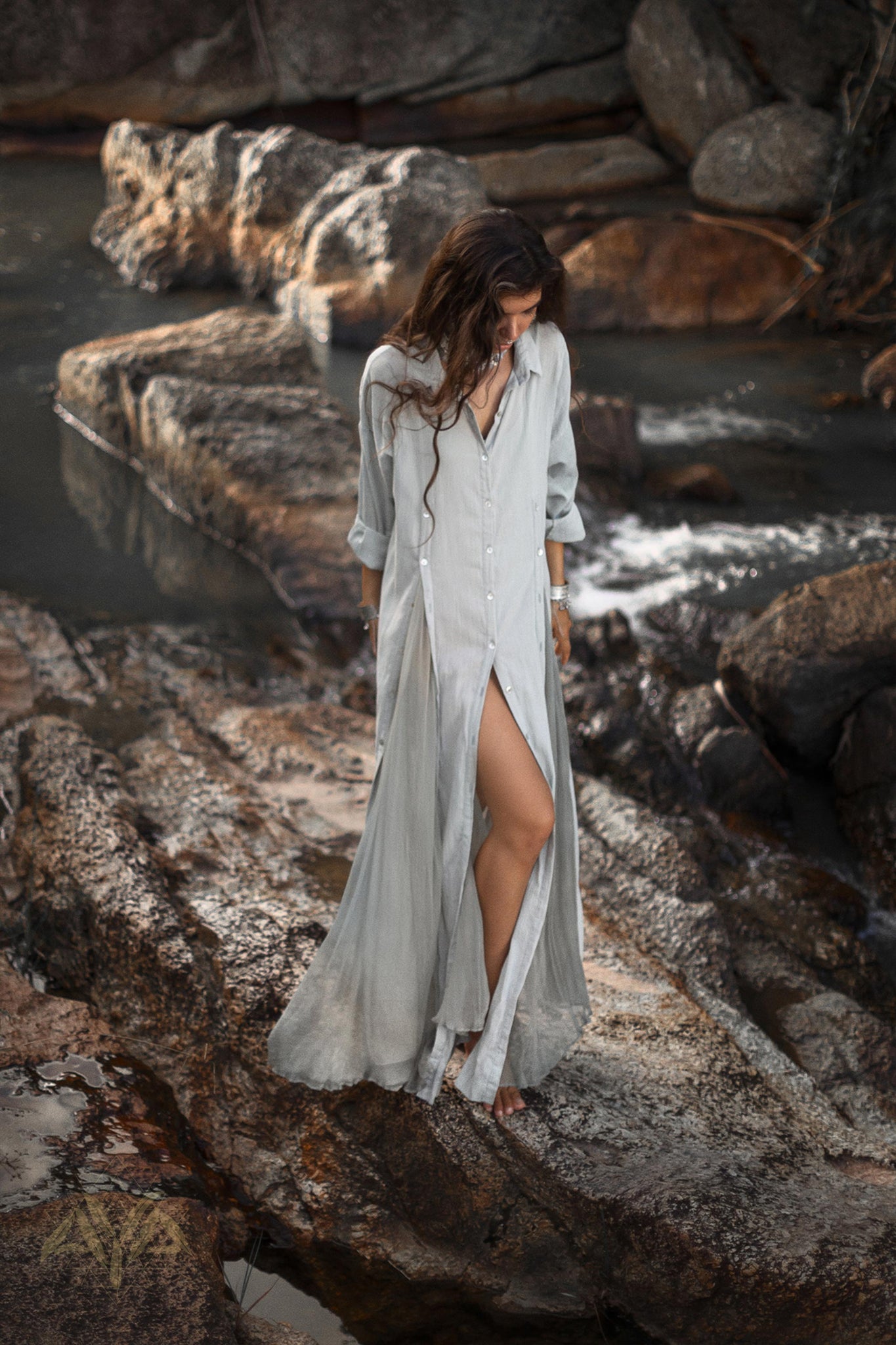 A woman in a light, long Powder Blue Kannika Shirt Dress by AYA Sacred Wear stands barefoot on rocky terrain near a stream. As she looks down, her loose hair cascades past her shoulders. The backdrop of flowing water and rocks enhances the serene, natural environment.