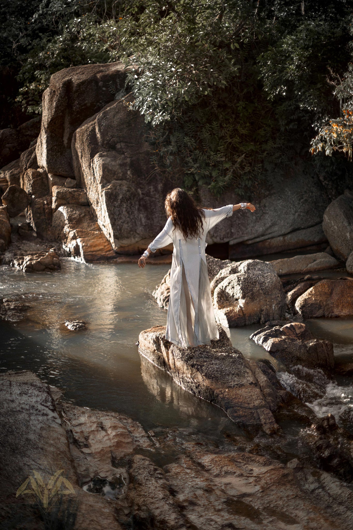 A person with long hair stands on a rock in the middle of a stream, surrounded by large boulders and lush greenery. They are wearing AYA Sacred Wear's Powder Blue Kannika Shirt Dress, a multiway button-down dress and wrap cover-up made from organic crinkle cotton and botanical dyes. Their arms are outstretched, appearing to balance or pose gracefully as sunlight filters through the trees.