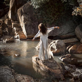 A person with long hair, clad in an organic cotton Powder Blue Kannika Shirt Dress by AYA Sacred Wear, stands on a rock in a tranquil stream. Surrounded by large stones and lush greenery, the scene exudes serenity enhanced by botanical dyes.