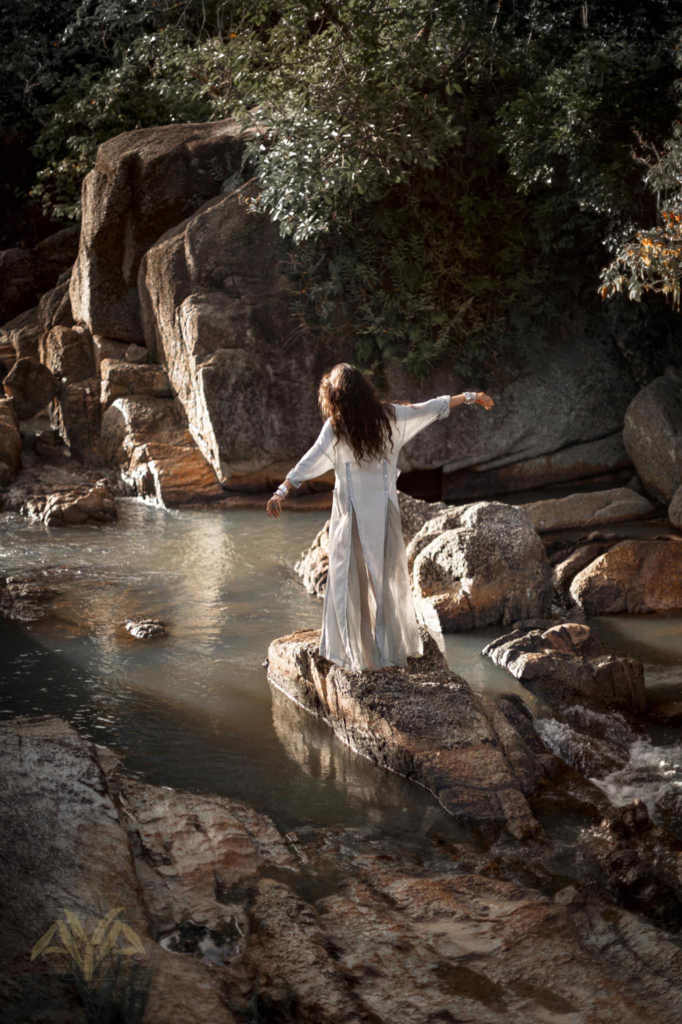 A person with long hair, clad in an organic cotton Powder Blue Kannika Shirt Dress by AYA Sacred Wear, stands on a rock in a tranquil stream. Surrounded by large stones and lush greenery, the scene exudes serenity enhanced by botanical dyes.
