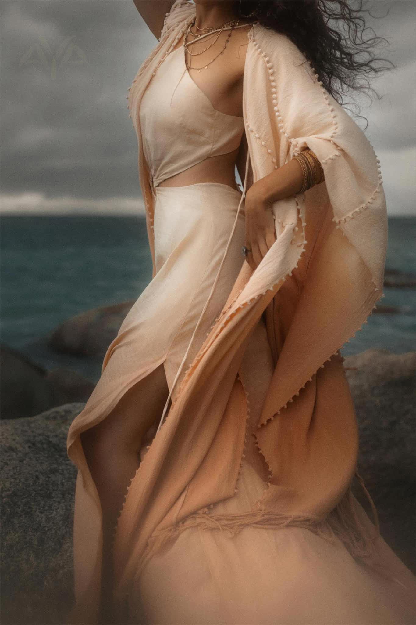 Standing on rocky sea terrain, a person wears the AYA Sacred Wear Powder Pink Boho Poncho, an elegant handmade bohemian cape with textured edges. Layered necklaces and bracelets add charm against the ocean and moody sky backdrop.