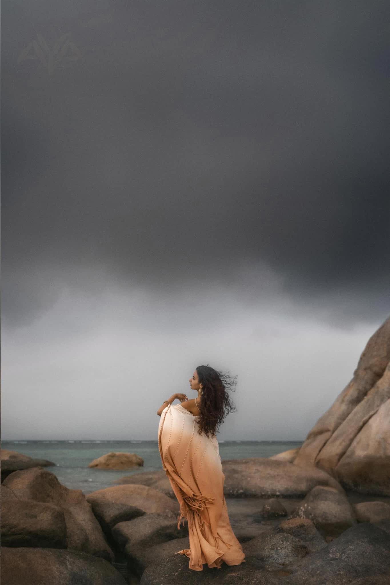 A woman in a flowing Powder Pink Boho Poncho by AYA Sacred Wear stands on rocky terrain by the sea. Stormy gray clouds loom overhead, and her long hair blows in the wind, enhancing the dramatic atmosphere.