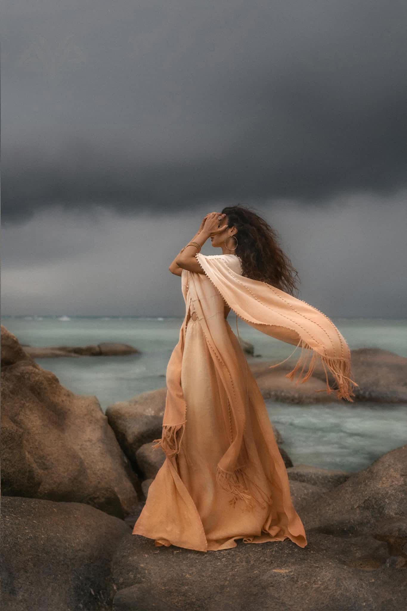 A woman stands on rocks by the sea in AYA Sacred Wear's Powder Pink Boho Poncho, a flowing ombre sustainable mantel. Her hair blows as she gazes at the stormy sky, with dark clouds and a turbulent ocean in the background.