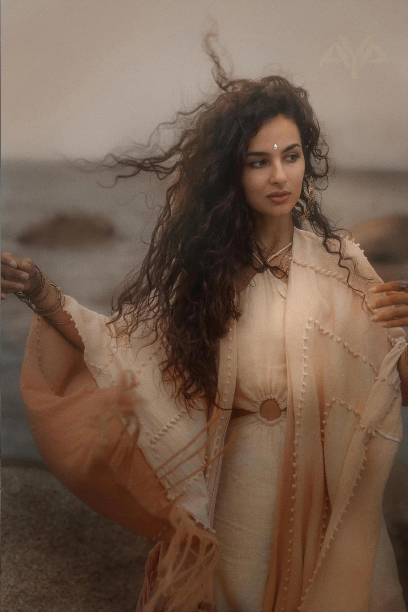 A woman with curly hair stands by the sea in a Powder Pink Boho Poncho from AYA Sacred Wear. The wind gently stirs her shawl. Calm waters and rocks lie under a muted sky, reflecting the ombre tones of this sustainable wrap achieved through botanical dyeing.