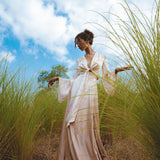 Model wearing a Powder Pink Venus Peace Silk Skirt, a minimalist wrap skirt made from organic materials, standing gracefully in a field of tall grass under a bright blue sky. The skirt flows elegantly as she poses with her arms raised.