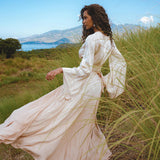 model wearing a Powder Pink Venus Peace Silk Skirt, showcasing a minimalist wrap skirt design, standing in a lush green field with mountains and a body of water in the background. The skirt flows elegantly with a gentle breeze, highlighting its organic summer aesthetic.