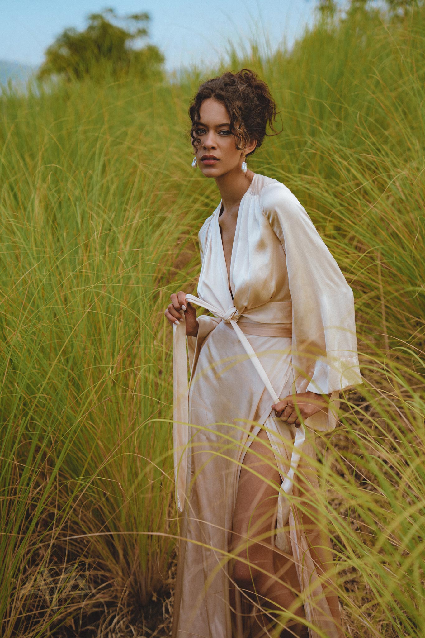 A model stands among tall green grass, wearing a flowing powder pink silk wrap skirt. The outfit features minimalistic design elements with a tied waist and long sleeves. The scene captures a serene summer vibe, emphasizing the organic nature of the fabric and style. The model gazes thoughtfully at the camera, contrasting with the lush landscape.