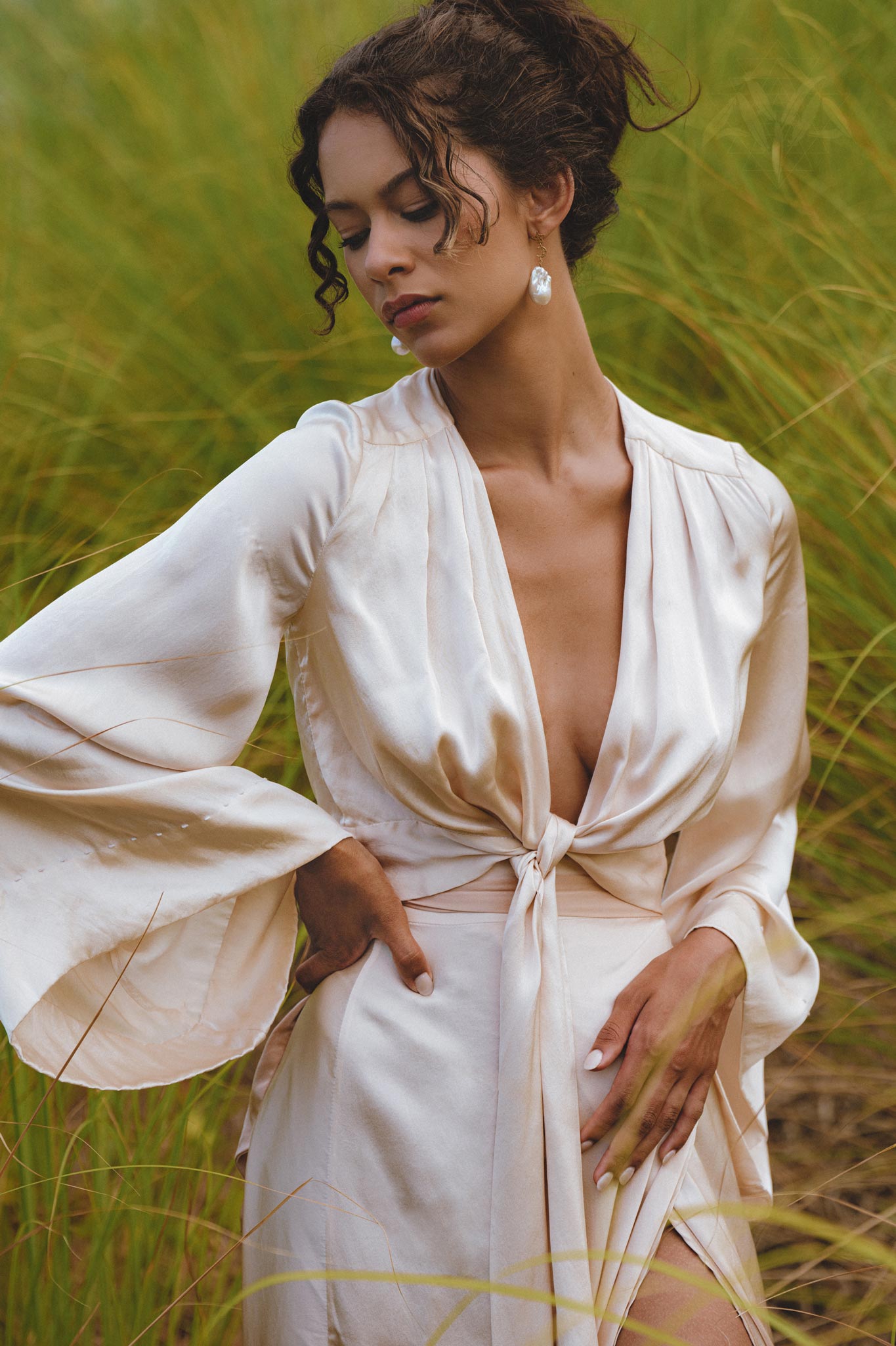 A woman poses gracefully in a lush green setting, wearing a powder pink silk wrap skirt. The skirt features a minimalist design, elegantly tied at the waist and complemented by a matching top with flowing sleeves. She has a serene expression and is adorned with statement earrings, adding a touch of sophistication to the organic summer look.